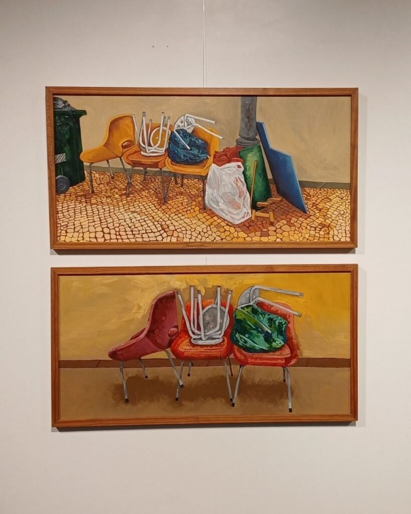 Duas pinturas a óleo de Marta Facco, exibidas na exposição Obsolescência, emolduradas uma sobre a outra na parede. Na de cima, uma cadeira laranja e outras cadeiras empilhadas de pernas para o ar sobre calçada de pedras portuguesas, ao lado de uma lixeira verde, uma sacola plástica branca amarrada, um vaso azul e uma placa de metal apoiada na parede. Na de baixo, três cadeiras vermelhas em fileira, com cadeiras de metal empilhadas de cabeça para baixo sobre os assentos e um saco verde escuro sobre uma delas, contra um fundo amarelo e piso marrom.