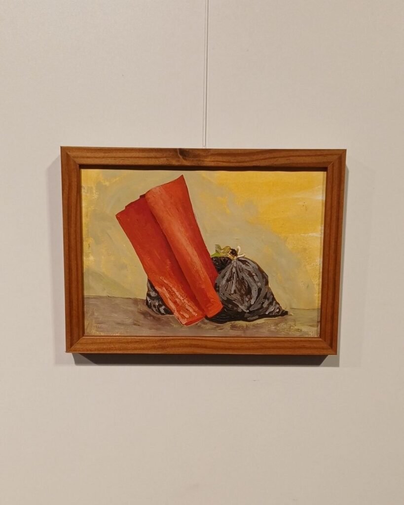 Pintura a óleo de Marta Facco na exposição Obsolescência, emoldurada em madeira, com dois rolos de tapete ou carpete laranja apoiados de pé lado a lado, encostados numa parede, ao lado de um saco de lixo preto amarrado com uma flor branca presa no nó. Fundo em tons de amarelo e cinza, cena montada sobre uma superfície acinzentada.
