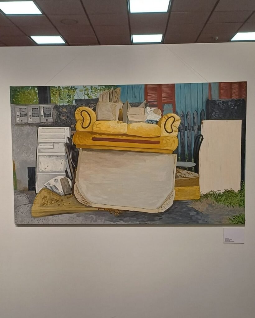 Pintura a óleo de grande formato de Marta Facco, parte da exposição Obsolescência, numa parede de galeria sob teto com luminárias quadradas. A cena retrata um sofá amarelo virado de cabeça para baixo, com o encosto branco apoiado no chão de uma rua, cercado de móveis descartados: um armário branco, um ferro de passar velho, caixas de papelão e sacolas empilhadas em cima do sofá, e uma placa de madeira clara encostada ao lado. Ao fundo, um portão azul com detalhes vermelhos, uma cerca de madeira e vegetação verde.