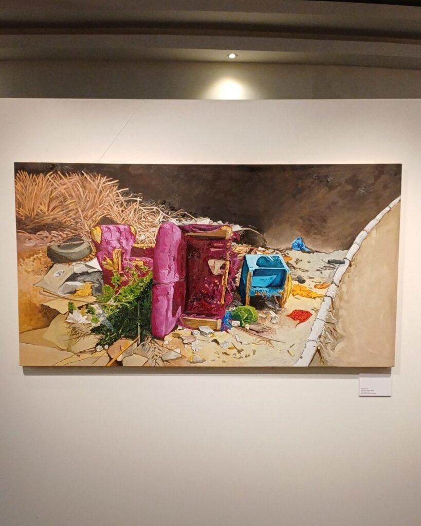 Pintura a óleo em formato panorâmico de Marta Facco, na exposição Obsolescência, iluminada por spot de teto na galeria. A cena mostra móveis estofados descartados à beira de uma calçada com meio-fio branco: uma poltrona rosa e um sofá vinho amontoados entre entulho, um berço ou cadeirinha infantil azul ao lado, cercados de vegetação seca, um pneu velho e fragmentos de gesso ou entulho de obra espalhados pelo chão de terra.
