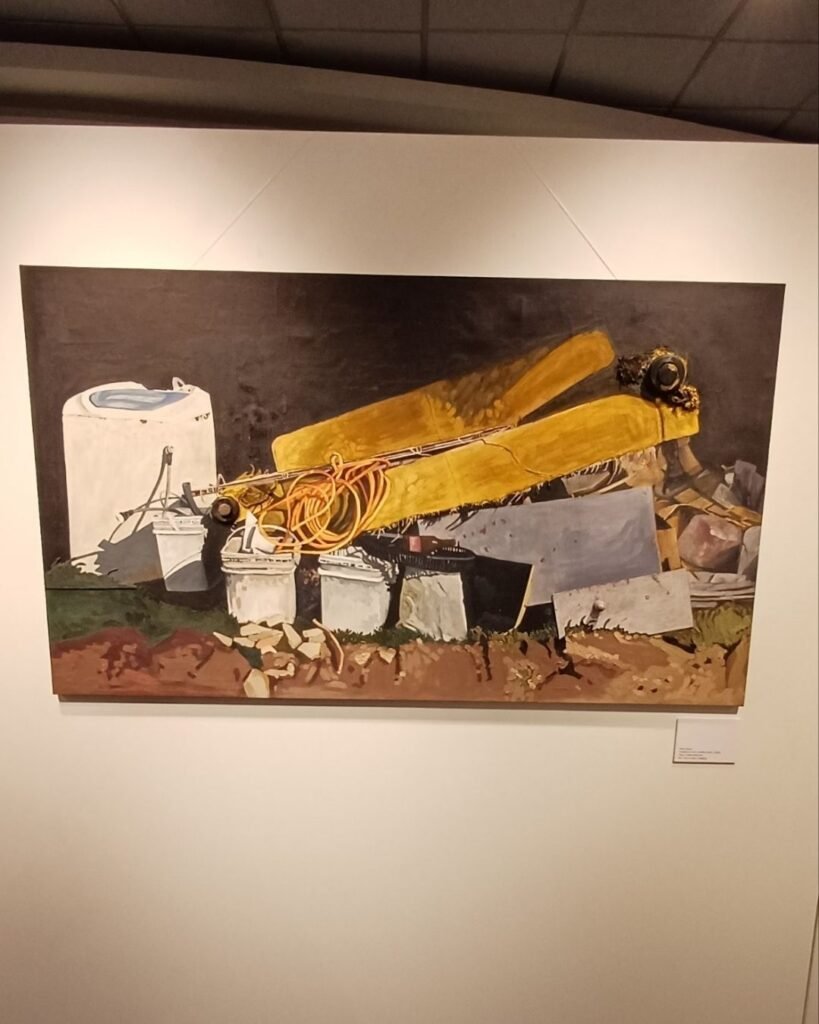 Pintura a óleo de Marta Facco, exibida na exposição Obsolescência, pendurada por fios na parede da galeria, com fundo escuro contrastando com os objetos descartados retratados. A cena mostra uma máquina de lavar roupas branca ao lado de baldes plásticos empilhados, uma mangueira laranja enrolada, dois colchões ou estofados amarelos dobrados um sobre o outro e entulho de construção espalhado sobre terra vermelha e vegetação rasteira.