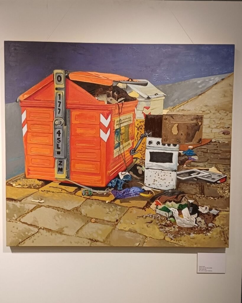 Pintura a óleo de grande formato de Marta Facco, na exposição Obsolescência, na parede da galeria, com placa de identificação da obra ao lado. A cena mostra uma caçamba de lixo laranja com faixas reflexivas brancas e o código "0 177 45L ZM" impresso na lateral, transbordando de entulho, ao lado de um fogão branco velho, um pequeno armário enferrujado e sacolas plásticas espalhadas sobre calçada de pedras, contra um céu azul ao fundo.