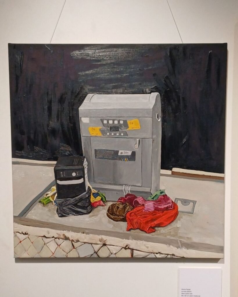 Pintura a óleo de Marta Facco, exibida na exposição Obsolescência, emoldurada em fundo escuro, pendurada por fios na parede da galeria, com placa de identificação da obra ao lado. A cena retrata um caixa eletrônico ou terminal de autoatendimento cinza descartado sobre uma calçada, ao lado de um gabinete de computador preto amarrado com fita adesiva, tecidos coloridos amassados em tons de vermelho, rosa e verde, e uma sacola de papel marrom, tudo sobre uma base de concreto com uma grade metálica ao fundo.