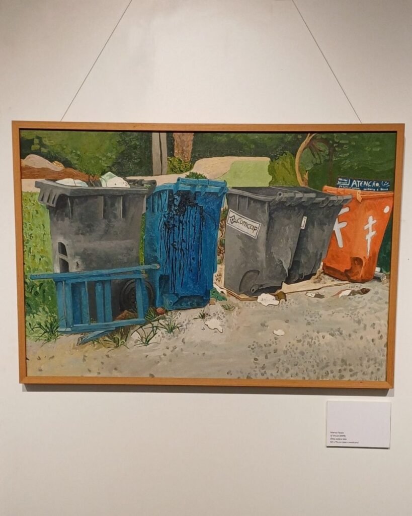Pintura a óleo de Marta Facco, na exposição Obsolescência, emoldurada, pendurada por fios na parede da galeria, com placa de identificação e vegetação verde ao fundo. A cena mostra uma fileira de lixeiras públicas danificadas: uma cinza aberta e amassada, uma azul rasgada e caída sobre um carrinho de mão, duas cinzas com a marca "Comcap" (empresa de limpeza urbana de Florianópolis) e uma laranja com adesivos, todas apoiadas sobre uma calçada de terra e cascalho, com uma placa de "atenção" ao fundo.