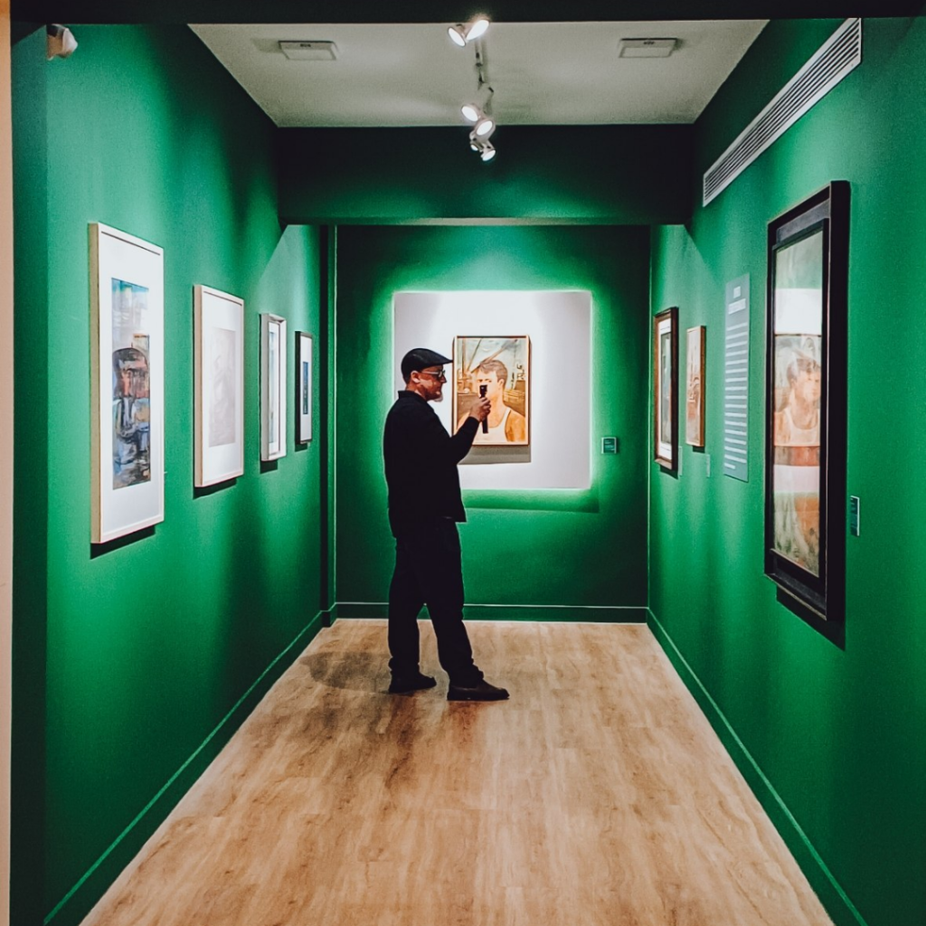 Marco filmando para o canal Onde Ver Arte na sala verde da exposição Diários Afetivos, no Instituto Collaço Paulo, com telas emolduradas nas paredes ao redor
