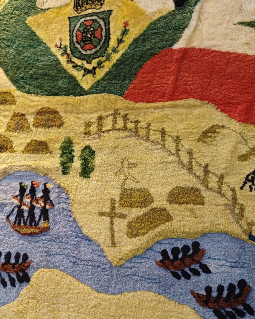 Detalhe do mapa no painel central: terra amarelada com morros, cruz e uma linha de cerca, cercada de água azul onde navegam um navio de três mastros e barcos a remo com marinheiros. No alto aparece o brasão do Império.