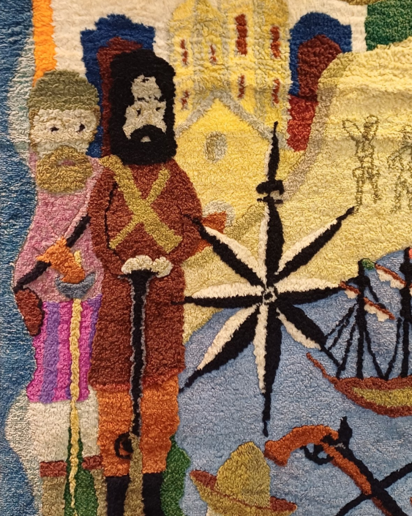 Detalhe do canto esquerdo do painel central: um homem barbudo de chapéu preto, casaco marrom e faixas amarelas cruzadas no peito, apoiado num bastão, ao lado de outro homem de barba, túnica rosa e machado. À frente deles estão uma rosa dos ventos e um navio de velas, com uma igreja amarela ao fundo.