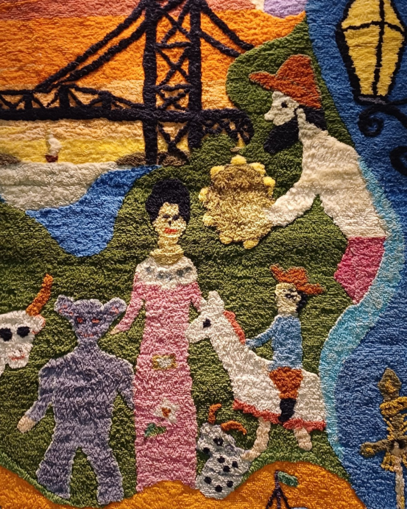 Detalhe do painel da esquerda: a Ponte Hercílio Luz em fios escuros contra um céu de pôr do sol laranja, uma mulher de vestido rosa e cabelos pretos, uma figura cinza-azulada de orelhas redondas, um boi branco de chifres, um menino sobre um cavalinho branco e um homem de chapéu vermelho.