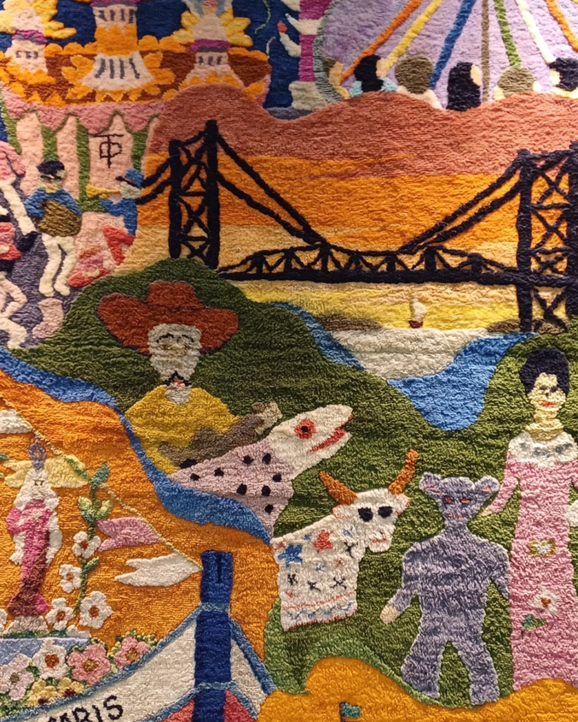 Detalhe do painel da esquerda com a Ponte Hercílio Luz ao pôr do sol, e embaixo as figuras da brincadeira: boi de cabeça branca, bicho de bolinhas e a figura cinza-azulada ao lado da mulher de rosa. No canto inferior esquerdo, parte de um barco com flores e uma santa, e o nome do barco cortado, “...ARIS”.