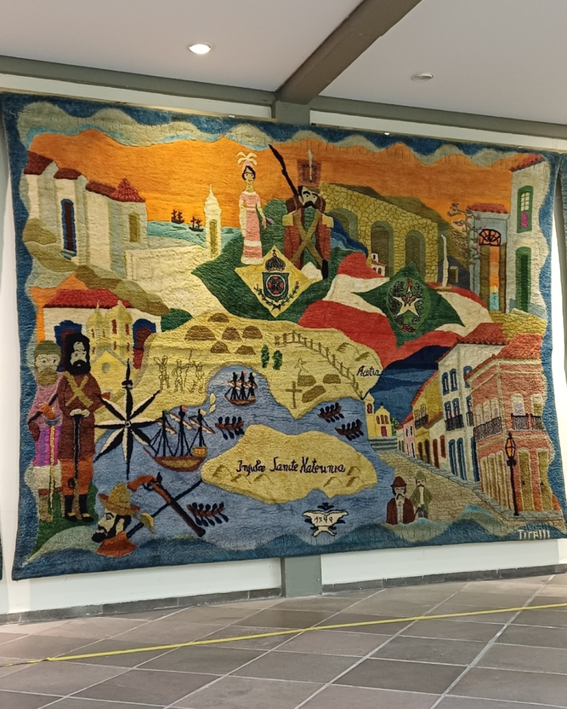 Painel central da tapeçaria Almir Tirelli pendurado sob o teto com focos de luz. O mapa antigo da Ilha, cercado de caravelas e barcos a remo, ocupa o centro, com bandeiras, brasão, arcadas de pedra, sobrados antigos e a assinatura “Tirelli” no canto inferior direito.