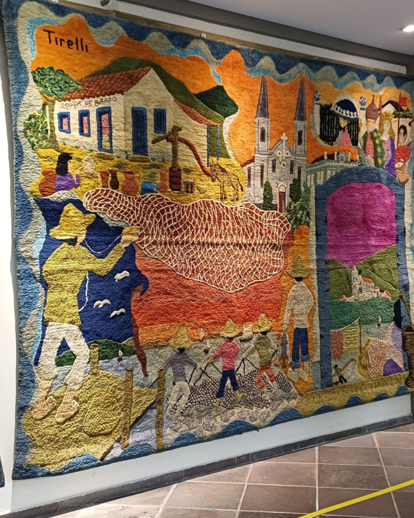 Painel da direita da tapeçaria, visto em ângulo numa parede de museu, com um pescador de chapéu amarelo lançando uma grande tarrafa sobre o mar azul, casa branca com a inscrição “Louça de Barro”, igreja de duas torres, arco rosa com paisagem ao fundo e homens puxando a rede na praia. A assinatura “Tirelli” aparece no canto superior esquerdo.