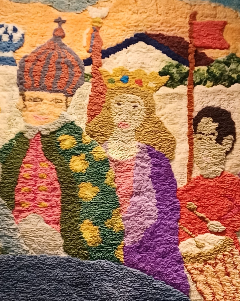 Detalhe do cortejo no canto superior direito do painel da direita: uma figura de coroa listrada em forma de cúpula e casaco verde com bolas amarelas, ao lado de uma figura de coroa dourada, cabelos loiros e manto roxo, e de um homem de vermelho com baqueta de tambor, sob um estandarte vermelho. Os nós de lã formam uma superfície bem texturizada.