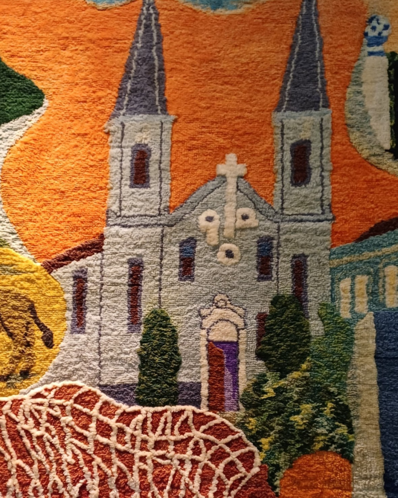 Detalhe de uma igreja de duas torres com pontas azul-acinzentadas, cruz branca na fachada e cipreste dos dois lados, sob céu laranja. Na parte inferior aparece a trama vermelha e branca da rede de pesca.