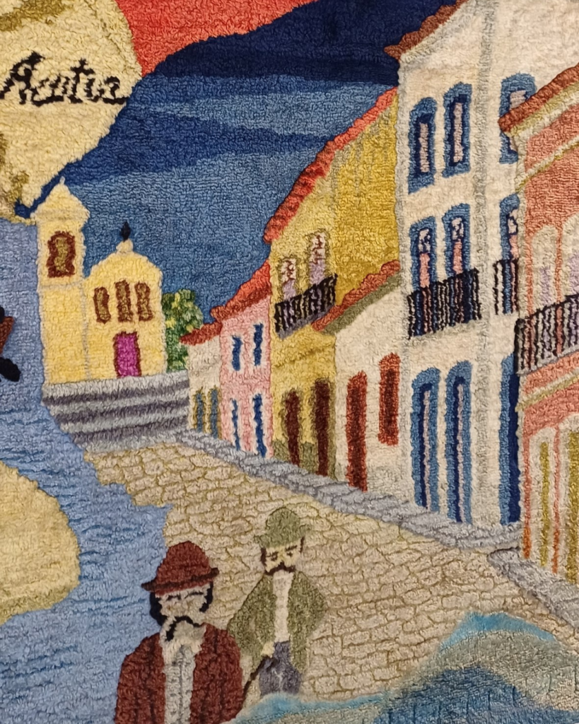 Detalhe do painel central: rua de pedras ladeada por sobrados coloridos com sacadas de ferro, uma igreja amarela com porta rosa junto à água e, em primeiro plano, dois homens de chapéu e bigode.
