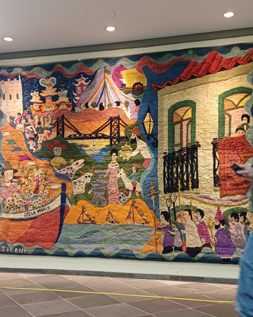 Painel da esquerda da tapeçaria Almir Tirelli, na parede de um museu: a Ponte Hercílio Luz ao centro, o barco Stela Maris cheio de flores à esquerda, as figuras do boi de mamão sobre um morro verde, veleiros no mar e, à direita, um casarão de sacadas de ferro com a procissão em roxo e rosa. Um visitante aparece parcialmente na borda direita.
