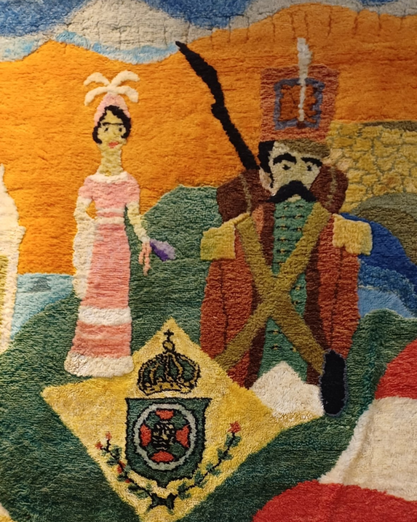 Detalhe do casal do painel central: mulher de vestido rosa longo, cabelos pretos e adereço branco de plumas, ao lado de um homem de bigode, chapéu alto vermelho com pluma e faixas verde-oliva cruzadas no peito. Abaixo deles está o brasão imperial com coroa sobre um losango amarelo.