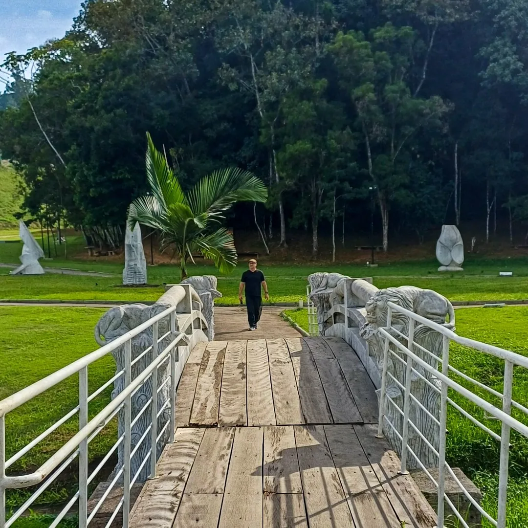 Imagem 03 Marco caminhando em direção à ponte de madeira do Parque das Esculturas Ilse Teske, em Brusque (SC), com esculturas em pedra no gramado
